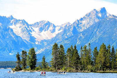 People canoeing on Jackson Lake with forested shoreline and snow-streaked Teton Range peaks in Grand Teton National Park, Wyoming, USA.