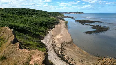 Aerial view of Cornelian Bay near Scarborough, showing a sandy beach, wooded coastal cliffs, exposed rocky reefs and the North Sea in North Yorkshire, England.