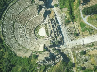 Overhead aerial view of the Great Theatre of Ephesus, showing its vast semicircular seating, stage complex, pathways, and surrounding ancient ruins.