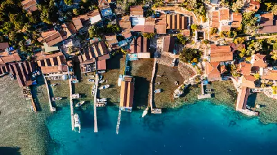Top-down aerial view of Kekova harbor, Antalya, Turkey, showing boats moored beside wooden piers, red-roofed coastal buildings, and clear turquoise water.