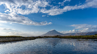Ol Doinyo Lengai volcano rising above a calm stream beneath a blue sky with scattered clouds in the Great Rift Valley near Lake Natron, northern Tanzania.