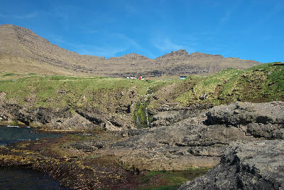 Rocky shoreline with small waterfall in the foreground and Viðareiði village beneath Villingardalsfjall mountain on Viðoy, Faroe Islands.