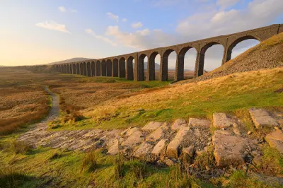 Ribblehead Viaduct illuminated by warm morning sunlight, with a winding stone footpath, limestone rocks and grassy moorland in the Yorkshire Dales, England.