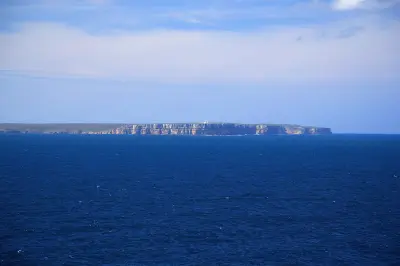Distant view across deep blue ocean toward the long sandstone cliffs of Point Perpendicular, with the lighthouse visible on the clifftop.