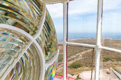 Close-up of the Cape Columbine Lighthouse Fresnel lens with views of the Atlantic Ocean and rocky coastline visible through the lighthouse lantern room windows near Paternoster, South Africa.