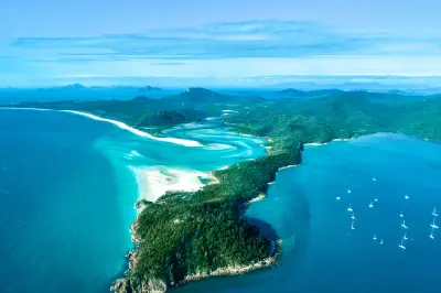 Wide aerial view of Whitehaven Beach, Hill Inlet, forested Whitsunday Island and yachts anchored in the surrounding blue water.
