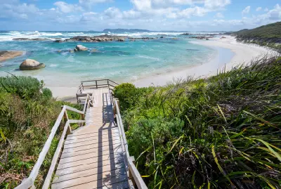 A wooden staircase descends a lush, grassy hill towards a curved white sand bay and clear turquoise water.