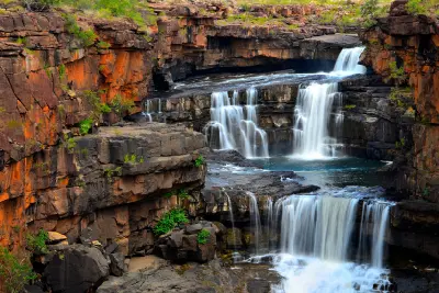 Multiple tiers of Mitchell Falls cascade over dark sandstone ledges between rugged orange cliffs and deep pools.