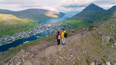 Hikers standing on a mountain viewpoint above Klaksvík with harbour, town and surrounding fjords visible on Borðoy, Faroe Islands.