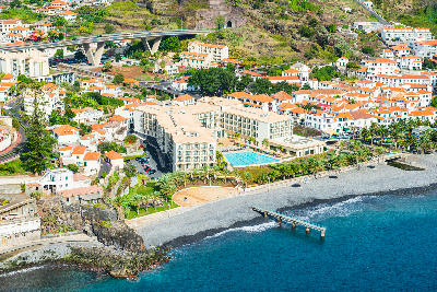 Aerial view of Santa Cruz in Madeira with coastal hotels, palm trees, pebble beach, and turquoise Atlantic Ocean along the shoreline.