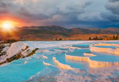 White travertine terraces filled with turquoise thermal water at Pamukkale, Turkey, illuminated by a dramatic sunset over the surrounding mountains.