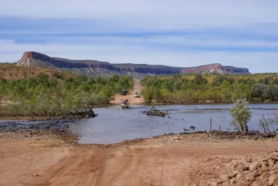 Four-wheel-drive vehicles cross the shallow Pentecost River with the red cliffs of the Cockburn Range rising in the background.