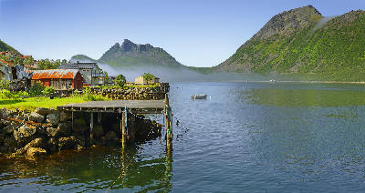 Gryllefjord village Senja Norway with wooden pier, houses and calm fjord water with mountains and low mist in background.