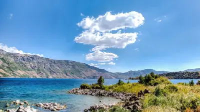 Turquoise Nemrut Crater Lake with a rocky shoreline and steep volcanic caldera walls beneath a bright blue sky.