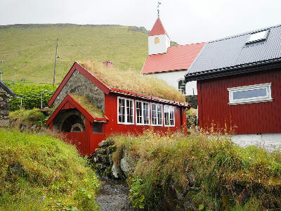 Red wooden watermill with grass roof and church in Mikladalur village on Kalsoy, Faroe Islands.