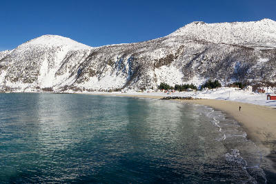Curving sandy beach at Bøvær on Senja with turquoise water, snow-covered mountains, and scattered houses along the shoreline