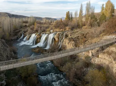 Aerial view of Muradiye Waterfall and a suspension footbridge crossing the river amid autumn trees in Van Province, Turkey.
