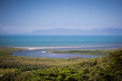 Sweeping vista from an elevated vantage point showing a winding coastal waterway, a sandy beach strip, and multi-shaded blue ocean water under a hazy sky.