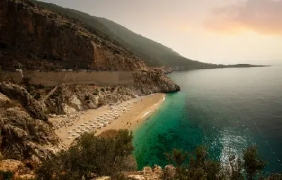 Elevated view of Kaputaş Beach, with rows of white umbrellas on a narrow sandy cove, clear turquoise Mediterranean water and a coastal road running along the steep rocky cliffs.