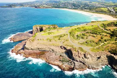 Aerial view across the rugged basalt cliffs and rock platforms of Bombo Headland, with waves breaking along the coast and Bombo Beach beyond.