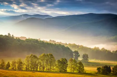 Golden morning light and mist hanging over the meadows and tree-lined valley of Cades Cove with blue Smoky Mountain ridges beyond.