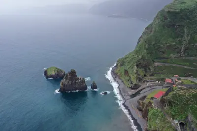 Volcanic sea stacks off the coast of Ribeira da Janela, with steep green cliffs and a narrow pebble shoreline along Madeira’s rugged north coast.
