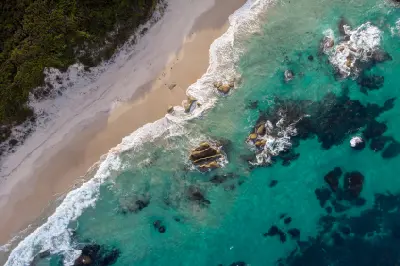 High-angle aerial photograph of a sandy beach meeting clear turquoise water and dark submerged reef rocks.