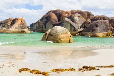  Large, rounded granite boulders rise from calm green-turquoise water next to a white sand beach with a lone seagull on the shore.