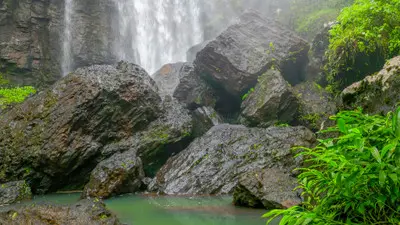 Large dark boulders and a pale turquoise pool at the base of Zillie Falls, with waterfall spray and green vegetation in the Atherton Tablelands.