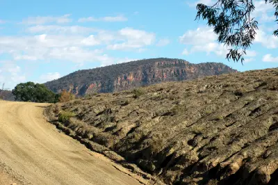 Gravel road beside eroded rock layers with distant mountain escarpments in Brachina Gorge, Flinders Ranges, South Australia.