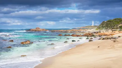 Cape Leeuwin Lighthouse seen beyond a sandy beach, turquoise water and granite rocks beneath dark storm clouds in Western Australia.