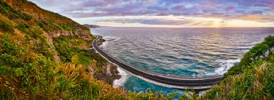Panoramic sunset view of Sea Cliff Bridge curving between steep coastal cliffs and the Tasman Sea, framed by lush vegetation.