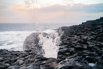 Seawater bursts upward from the Kiama Blowhole amid dark coastal rocks, with the Pacific Ocean and soft dawn sky beyond.