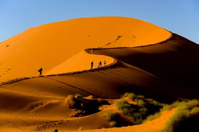People climbing sweeping orange sand dunes with curved shadowed ridges beneath a clear blue sky in the Sossusvlei area of Namibia.