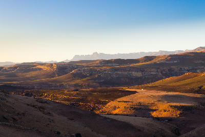 Panoramic view across the rolling valleys and sandstone escarpments of Golden Gate Highlands National Park, South Africa, with distant mountain ranges illuminated by warm evening light.