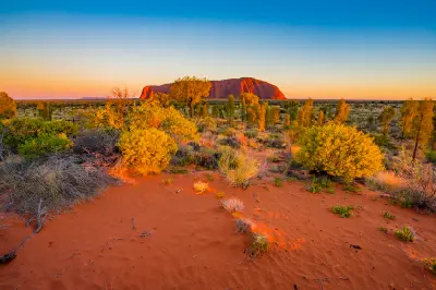 Uluru glowing in warm sunrise light above red sand and desert vegetation in Uluru-Kata Tjuta National Park, Australia.