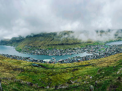 Wide elevated view of Klaksvík showing harbour, marina and dense town layout below steep terraced basalt mountains under low cloud on Borðoy, Faroe Islands.