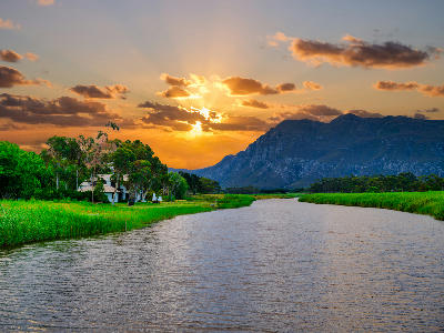 Golden sunset above the Klein River near Stanford in the Western Cape, with riverside houses, green reeds, and mountain silhouettes in the background.