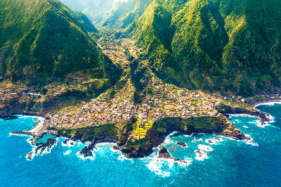 Aerial view of Seixal village in Madeira, showing steep green mountains, coastal terraces, and waves breaking against the volcanic coastline.