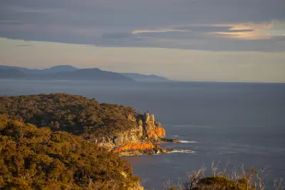 Orange-lichen-covered cliffs below Cape Tourville at sunrise, overlooking the Tasman Sea and distant coastline in Tasmania.