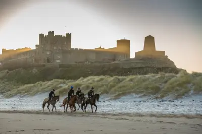 Four riders on horseback exercising along a snow-covered Bamburgh Beach at sunrise, with Bamburgh Castle rising above the dunes in Northumberland, England.