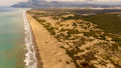 Aerial view along Patara Beach, showing an expansive golden sandy shoreline and dune system beside the Mediterranean Sea, backed by vegetation and rugged mountains.