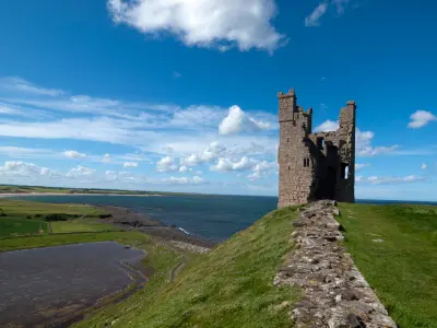 Ruined Liliburn Tower at Dunstanburgh Castle on a grassy coastal headland, with a surviving stone wall leading towards the tower and the North Sea stretching behind.