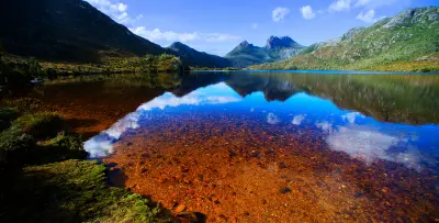 Cradle Mountain reflected in Dove Lake with clear water and a rocky orange lakebed in the foreground, Tasmania.