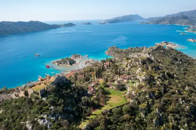 Elevated panoramic view from Kaleköy across the Kekova region, with historic castle walls, red-roofed village houses, rocky islands and vivid turquoise water stretching toward distant mountains.
