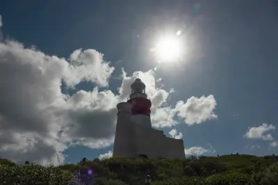 Cape Agulhas Lighthouse beneath bright sunlight and towering clouds above coastal fynbos in South Africa.