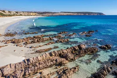 Elevated view of Stokes Bay on Kangaroo Island, showing layered coastal rocks, turquoise shallows, a sweeping white-sand beach and distant cliffs.