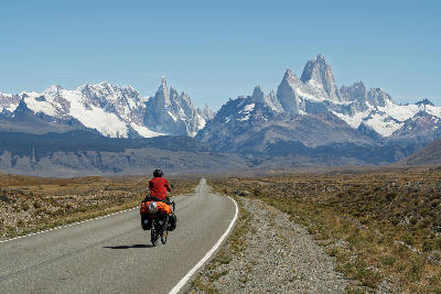 Cyclist riding a paved road across the Patagonian steppe toward the Fitz Roy massif near El Chaltén, Argentina.