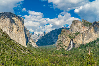 Yosemite Valley Tunnel View with El Capitan on left and Bridalveil Fall on right above dense pine forest under blue sky with clouds.