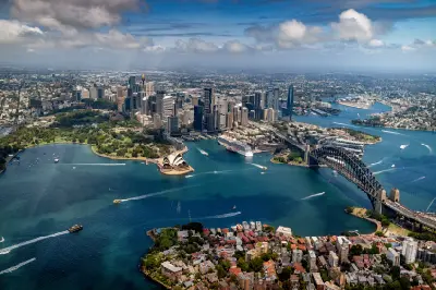 High aerial view across Sydney Harbour showing the Opera House, Harbour Bridge, CBD skyline, waterfront neighbourhoods and boats crossing the harbour.